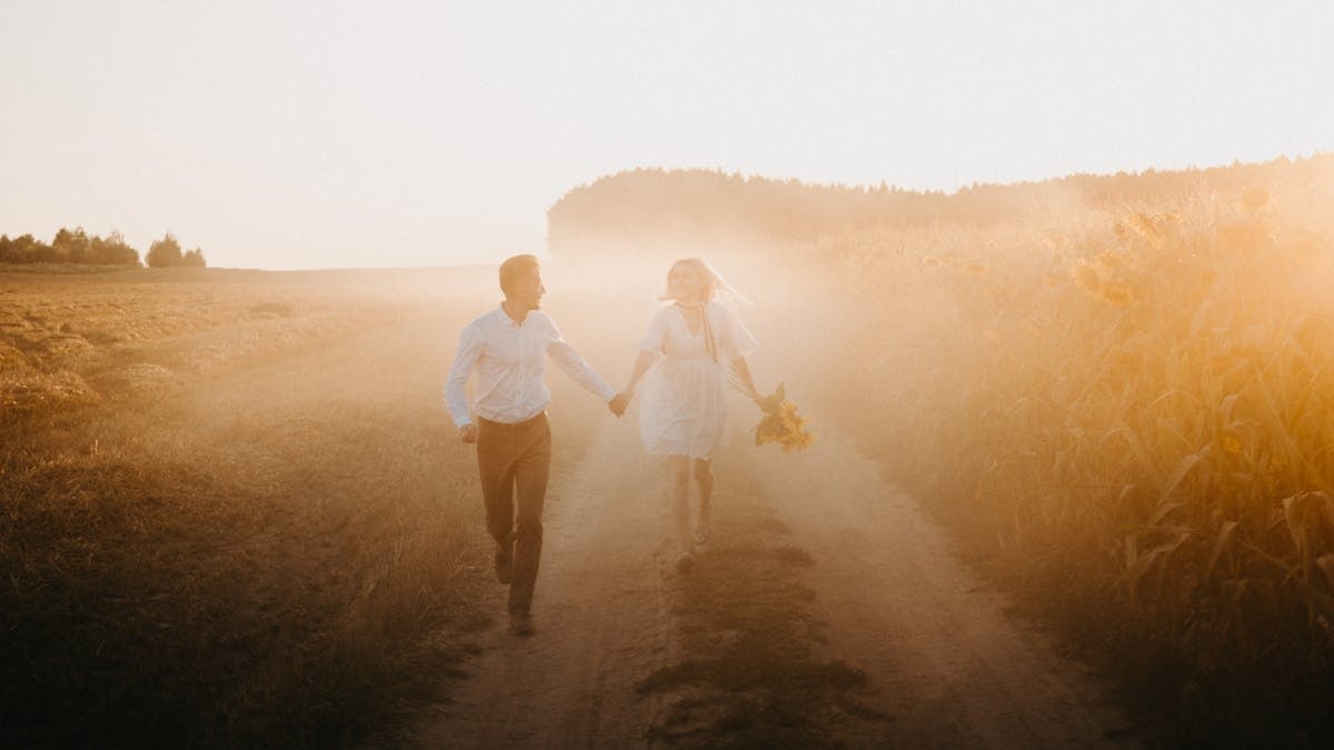 A couple holding hands and walking along a sunlit dirt road during sunset in a rural field.
